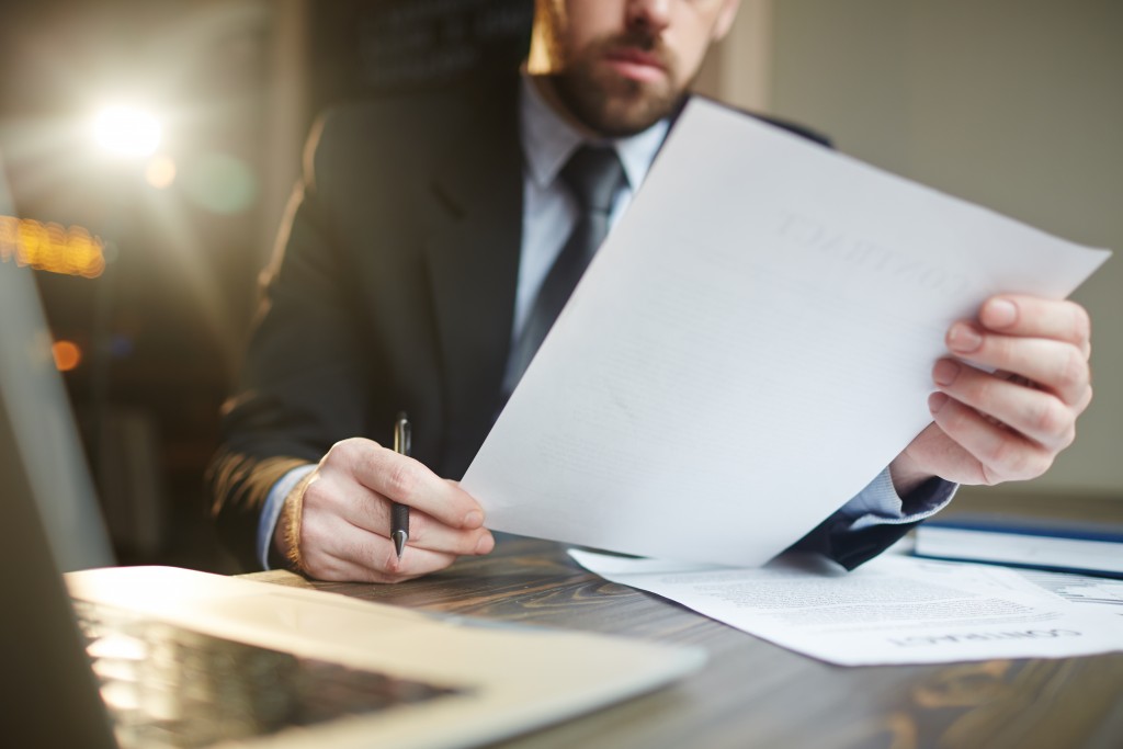 FBHA celebra entrada em vigor de lei que auxilia empresários durante a pandemia 1 Portrait of modern bearded businessman holding papers in hands, reading and analyzing contract documentation at desk with laptop