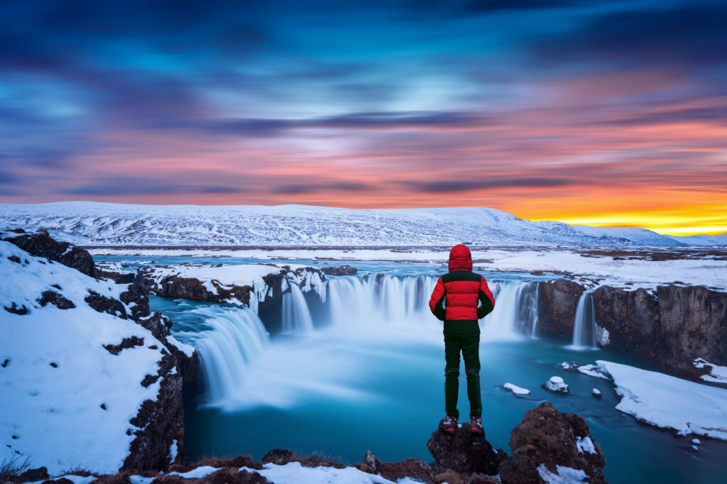 cachoeira de godafoss ao por sol no inverno islandia cara de jaqueta vermelha olha para cachoeira godafoss 30 destaques em 30 dias: confira as notícias mais importantes do Turismo em junho de 2025